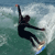 Pixelated figure in dark wetsuit riding a wave, arms extended, one hand raised, on a surfboard with blue and white details against a greenish ocean background.