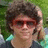 Pixelated static image of a person with dark curly hair wearing red-tinted sunglasses and a light-colored shirt against green foliage background.
