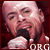 Pixelated close-up of a person with short light hair holding a microphone, wearing a dark shirt against a red background.