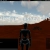 Pixelated static image shows a figure in dark clothing standing before a brown building under a cloudy sky.