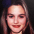 Pixelated close-up of a person with light brown hair, fair skin, and red lips smiling against a dark background.