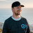 Pixelated image of a person wearing a dark cap and dark sweater with light blue collar, standing against a blurred background.