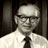 Pixelated grayscale portrait of a person wearing glasses, a light-colored shirt, and a dark tie against a dark background.
