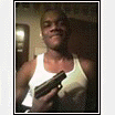 Static image of a person in a white tank top holding a black handgun, set against an indoor background with dim lighting and visible wall fixtures.