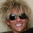 Pixelated close-up of a person with light brown hair wearing dark sunglasses and showing teeth in a smile against a neutral background.