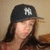 A person wearing a dark baseball cap with a white NY logo, long dark hair, and a red top against a light brown background.