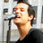 A pixelated static image shows a person with light skin and brown hair speaking into a microphone, wearing a dark shirt against a light background.