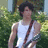 Pixelated image of a person in a white tank top with dark hair and a brown strap across the chest, standing outdoors against green foliage.