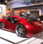 A red sports car with black accents is positioned on a white surface, viewed from a low angle with a blurred background featuring indistinct shapes and colors.