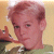Pixelated image of a person with light brown hair wearing a green shirt, holding a small object near their face against a beige background.
