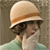 Pixelated image of a person wearing a light-colored hat with a brown band, dark sunglasses, and a light green top against a neutral background.