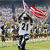 A pixelated sports scene shows a player in white jersey number 21 running on grass with an American flag waving behind, crowd visible.
