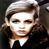 A static image of a person with short light brown hair styled in a side part, wearing a dark coat with a fur collar and a white shirt with a black tie.