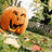 Pixelated orange pumpkin with carved face, white base, green foliage background, static image.
