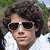 Pixelated close-up of a person with curly dark hair wearing white-framed sunglasses and a light-colored shirt against green foliage background.