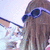 Pixelated image of a person with long brown hair wearing blue sunglasses, holding an object against a light background.