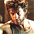 Pixelated image of a person with brown hair wearing dark clothing, holding an object near their face against a blurred background.