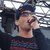 A person in a dark blue cap and striped sweater holds a microphone near their mouth against a blurred background with visible text overlay.
