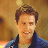Static pixelated image of a person with short brown hair wearing a dark blue jacket over a light shirt against a warm-toned gradient background.