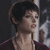 Pixelated close-up of a person with short dark hair wearing a dark purple garment, holding a white cylindrical object against a dark background.