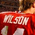 A red-shirted figure with light-colored hair holds a microphone, displaying 'ME 50M' text on the shirt against a dark background.