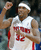 Pixelated image of a basketball player in white jersey with red number 12, raising right arm, wearing headband and dark background.