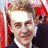 Pixelated static image of a person with light brown hair, wearing dark clothing and white collar, against red background elements.