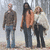 Three people stand outdoors in a blurred background; the central figure's right arm is raised and lowered