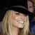 A pixelated close-up of a person with long blonde hair wearing a black hat, smiling with eyes closed against a dark background.