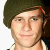 Pixelated close-up of a person with light skin wearing a dark green beret and white shirt, smiling slightly against a plain background.