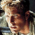 Pixelated image of a person with light brown hair tied back, wearing a light-colored shirt, and holding a dark object near their face.