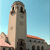 Pixelated static image of a tall, light-colored tower with dark rectangular windows against a clear blue sky, adjacent to a shorter building with a red-tiled roof.