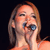Pixelated close-up of a person with light brown hair holding a microphone near their mouth against a dark background.