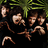 Pixelated image of multiple figures in dark clothing with green foliage background, some holding curved objects.