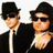 Two figures in black suits and fedoras pose together, one with an arm around the other's shoulder, against a white background.