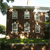 Static pixelated image of a brown house with multiple windows, green grass, and a tree on the left side under a light blue sky.