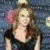 A woman with long brown hair wears a black top and blue jeans, standing on red carpet against blurred background.