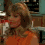 A woman with shoulder-length light brown hair wears an orange sleeveless top in a kitchen setting with teal cabinets and wooden shelves.