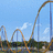 A roller coaster with orange tracks and blue support structures under a light blue sky with white clouds, viewed from ground level.