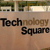 Static image with pixelated text reading "Technology Square" over a brown background, featuring a blurred top section with green and beige elements.