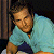Pixelated static image of a person in light blue polo shirt with dark collar and orange detail, leaning against wooden structure on dark background.