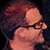 Pixelated close-up of a person's face with dark hair and glasses against a dark background, rendered in warm tones.