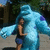 A blue, furry monster with purple spots stands beside a person in dark clothing and shorts on a paved area with trees.