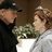 Two pixelated figures face each other; one wears a black cap and dark clothing, the other a light cap and light clothing, with a green plant-like object between them.