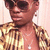 A person wearing a gold and brown striped shirt, dark sunglasses, and a silver necklace with a cross pendant appears in two distinct poses against a light pink background.