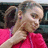 A pixelated close-up of a person with light brown hair wearing a bright pink top, hand raised near the face against a dark background.