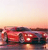 Pixelated static image of a red vehicle on a reflective surface under a gradient sky transitioning from light orange to pale pink.