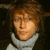 Pixelated close-up of a person with light brown hair and a white high-collared garment against a dark background.