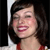 A pixelated close-up of a person with dark shoulder-length hair wearing a red top, smiling with visible teeth against a dark background.