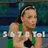 A pixelated image of a person in a green sleeveless top with dark hair tied back, standing against a blurred blue-green background with partial white text.